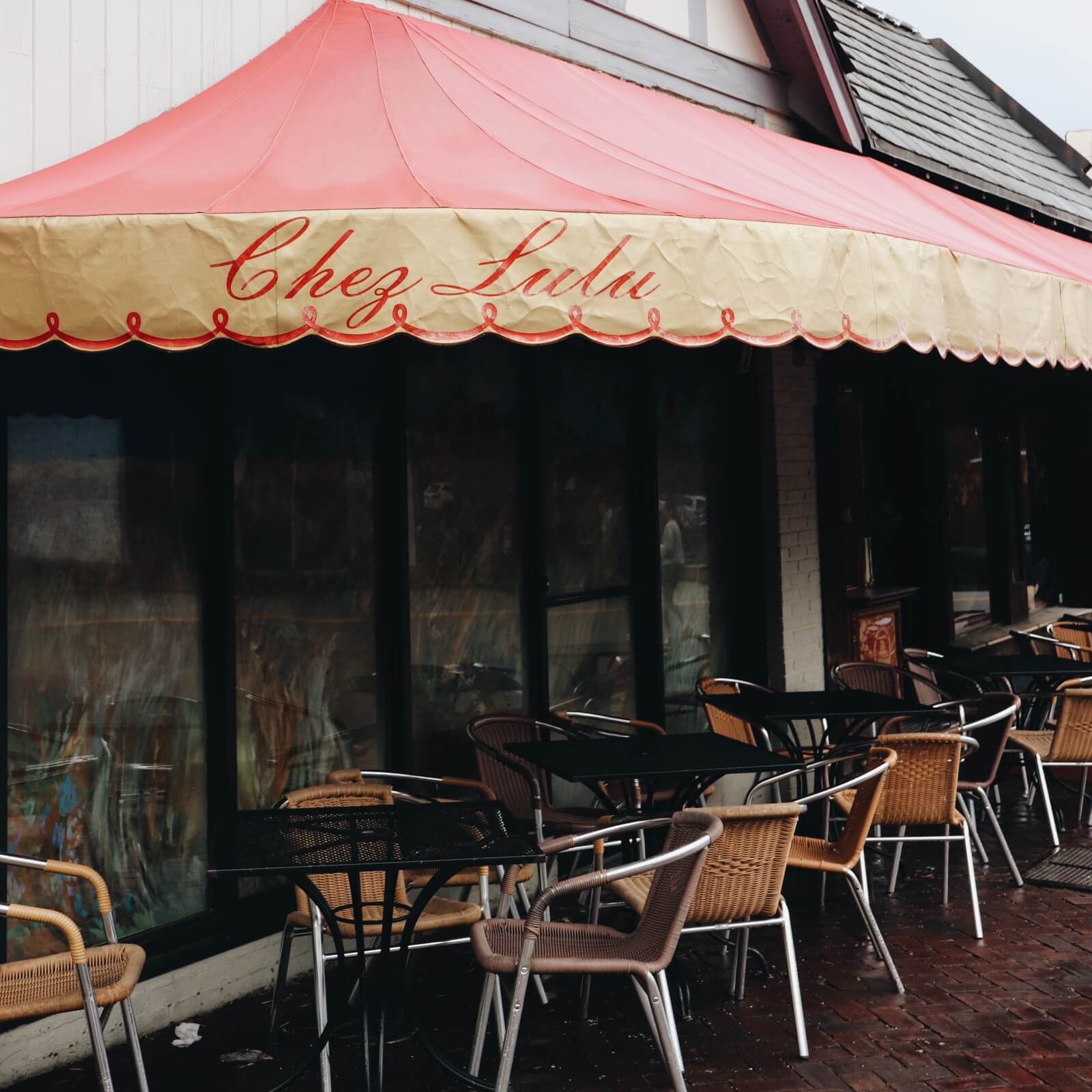 The outdoor seating area of a cafe in Birmingham features a red and yellow awning labeled "Chez Lulu." Empty tables and wicker chairs are arranged on the brick patio, perfect for enjoying afternoon tea.