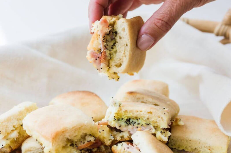 A hand holding a popular biscuit sandwich filled with salmon and seeds hovers above a pile of similar sandwiches on a cloth, capturing the culinary vibe of Birmingham in 2024.