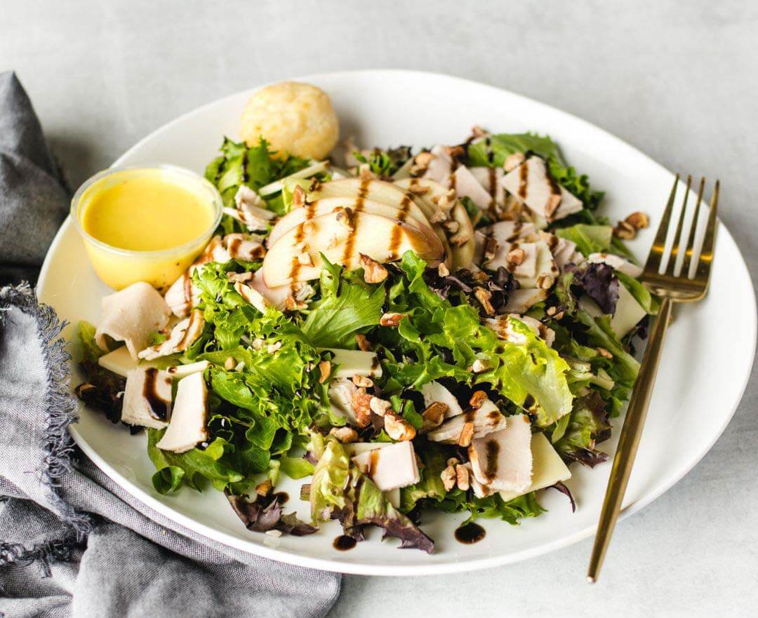 A salad with mixed greens, grilled chicken slices, and walnuts drizzled with dressing on a white plate.