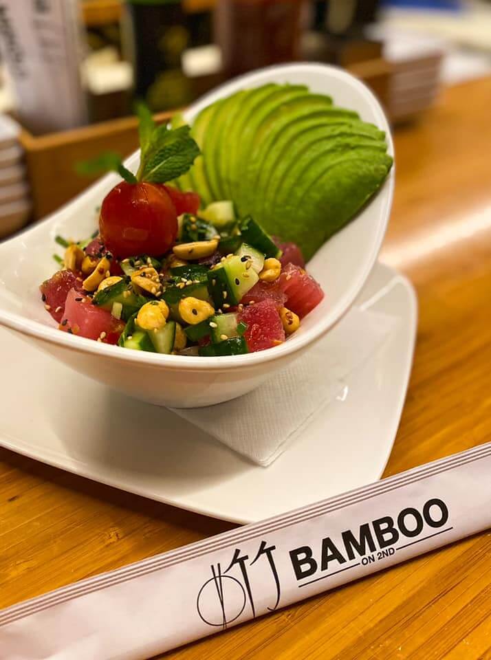 A bowl of salad with sliced avocado, tomatoes, cucumbers, nuts, and a mint garnish rests invitingly on a wooden table in Birmingham. Chopsticks labeled "Bamboo on 2nd" lie nearby, promising one of the best salads in the city.