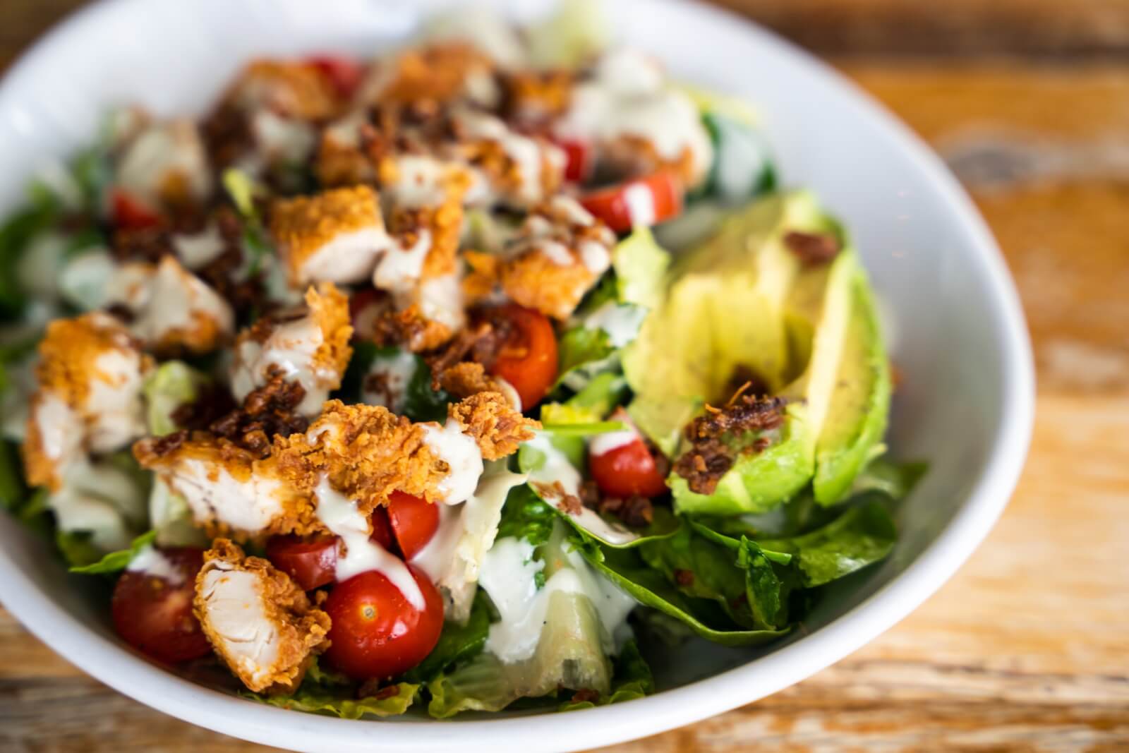 A bowl of one of Birmingham's best salads, featuring cherry tomatoes, sliced avocado, and fried chicken pieces drizzled with a creamy dressing, sits invitingly on a wooden table.