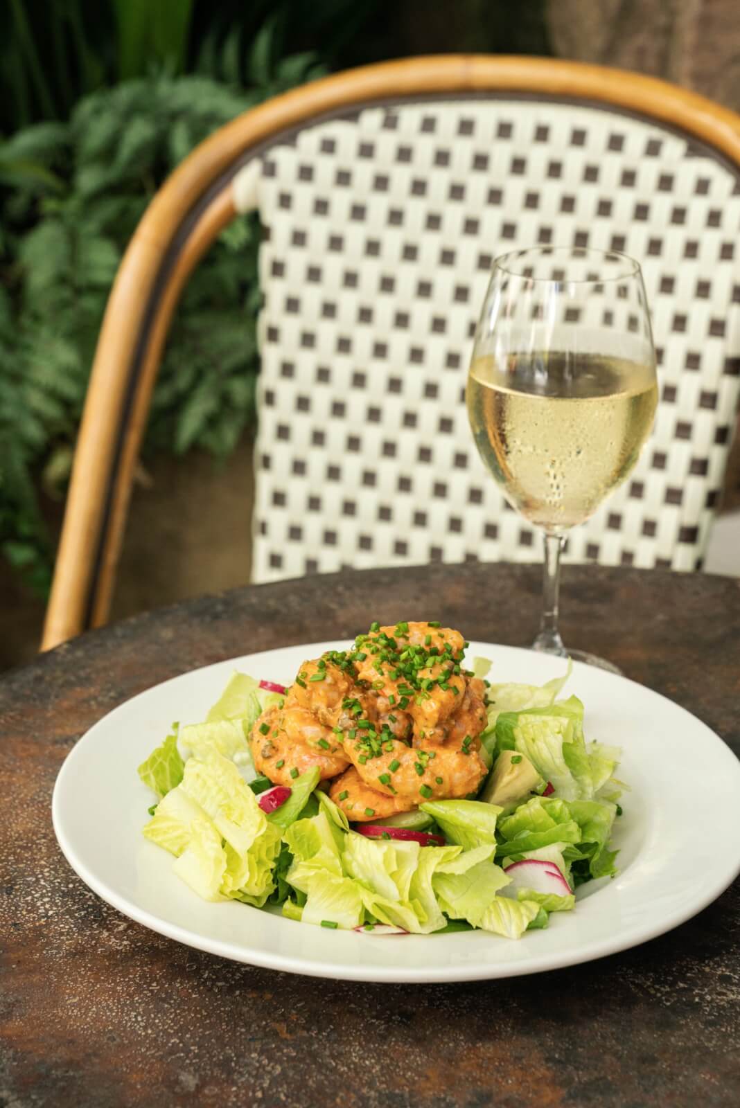 A plate of one of Birmingham's best salads featuring seasoned shrimp on a white dish, paired with a glass of white wine, sits invitingly on a wooden table with a woven chair in the background.