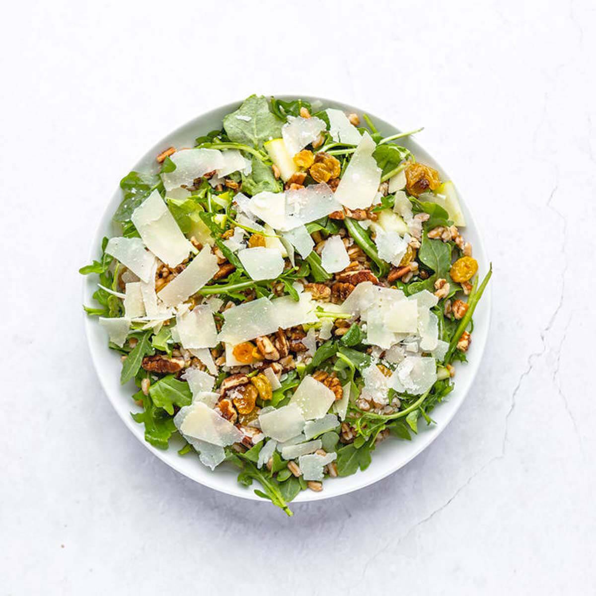 A bowl of salad with arugula, shaved Parmesan, walnuts, and golden raisins, all set against a crisp white background.