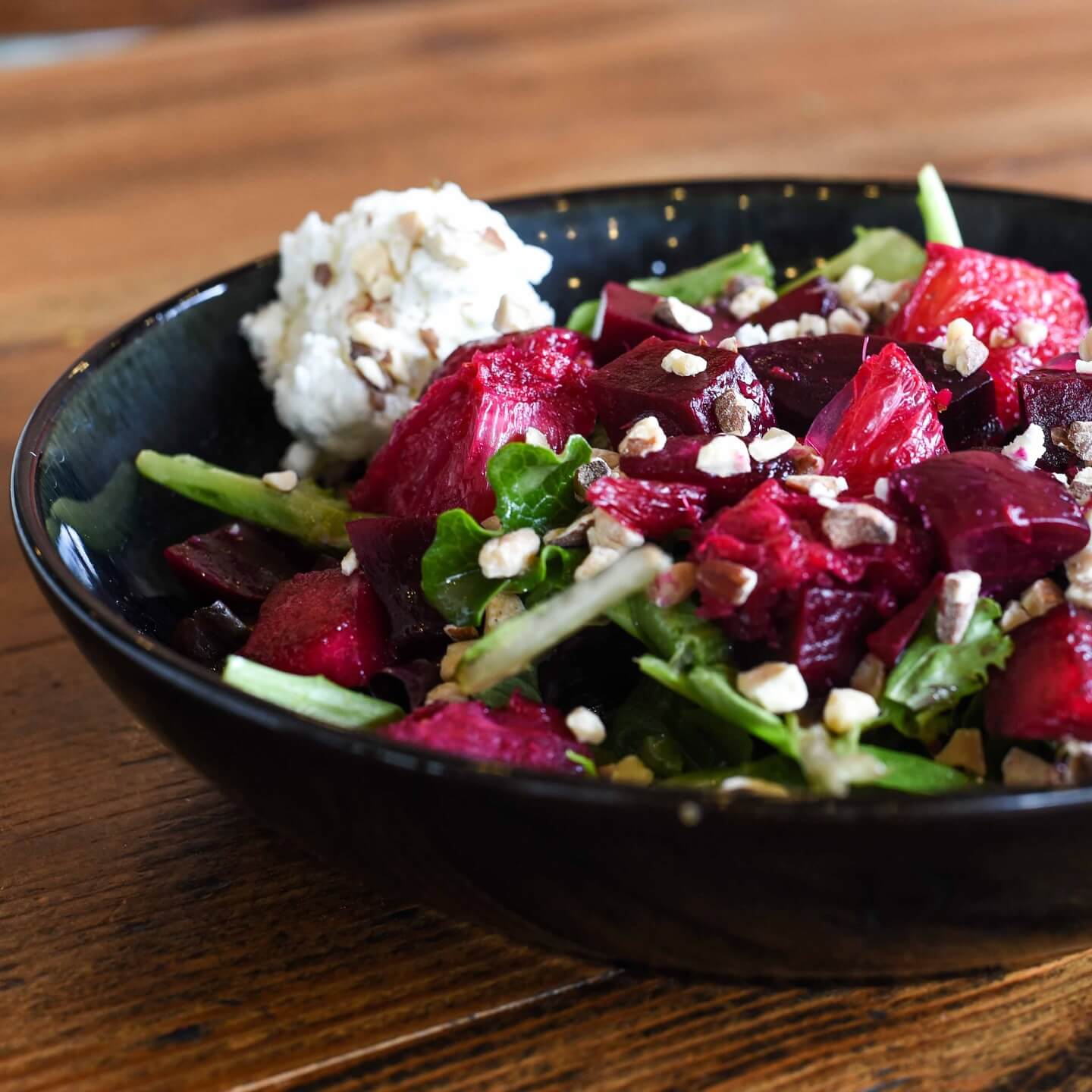 A black bowl contains one of Birmingham's best salads, featuring a beet and greens mix topped with chopped nuts and a dollop of white cheese, all elegantly presented on a wooden table.