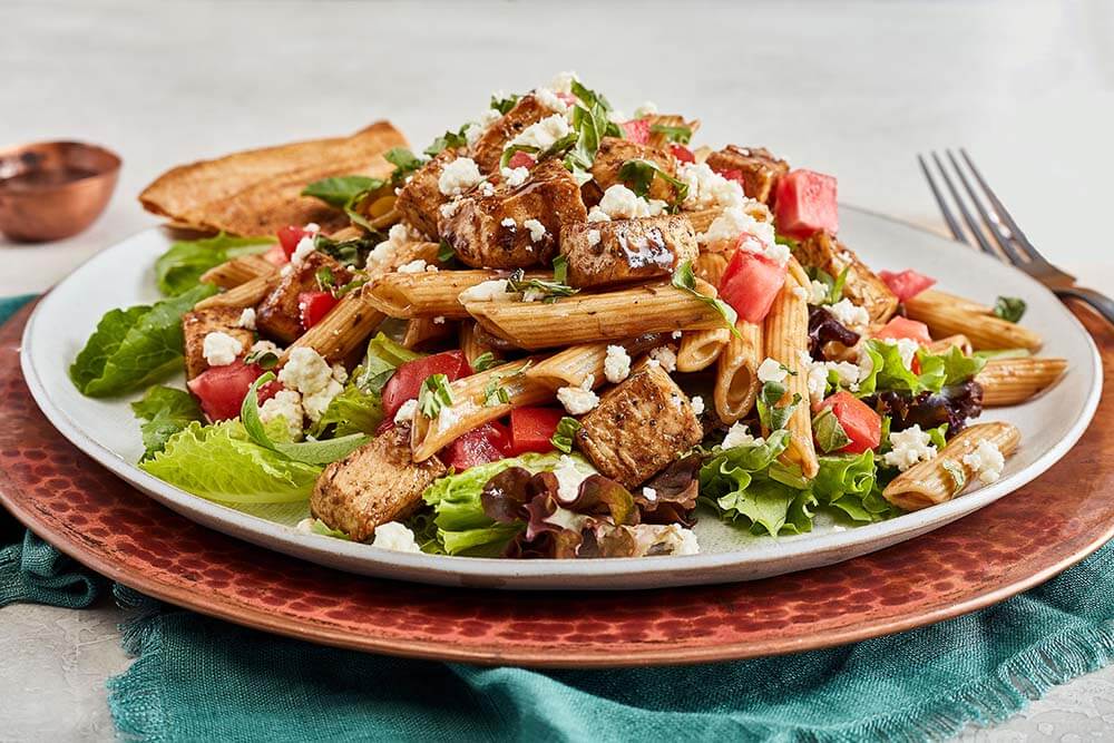 Savor one of Birmingham's best salads: a plate of pasta salad with grilled chicken, fresh lettuce, juicy tomatoes, feta cheese, and herbs, all served with a piece of bread on a copper-colored plate.