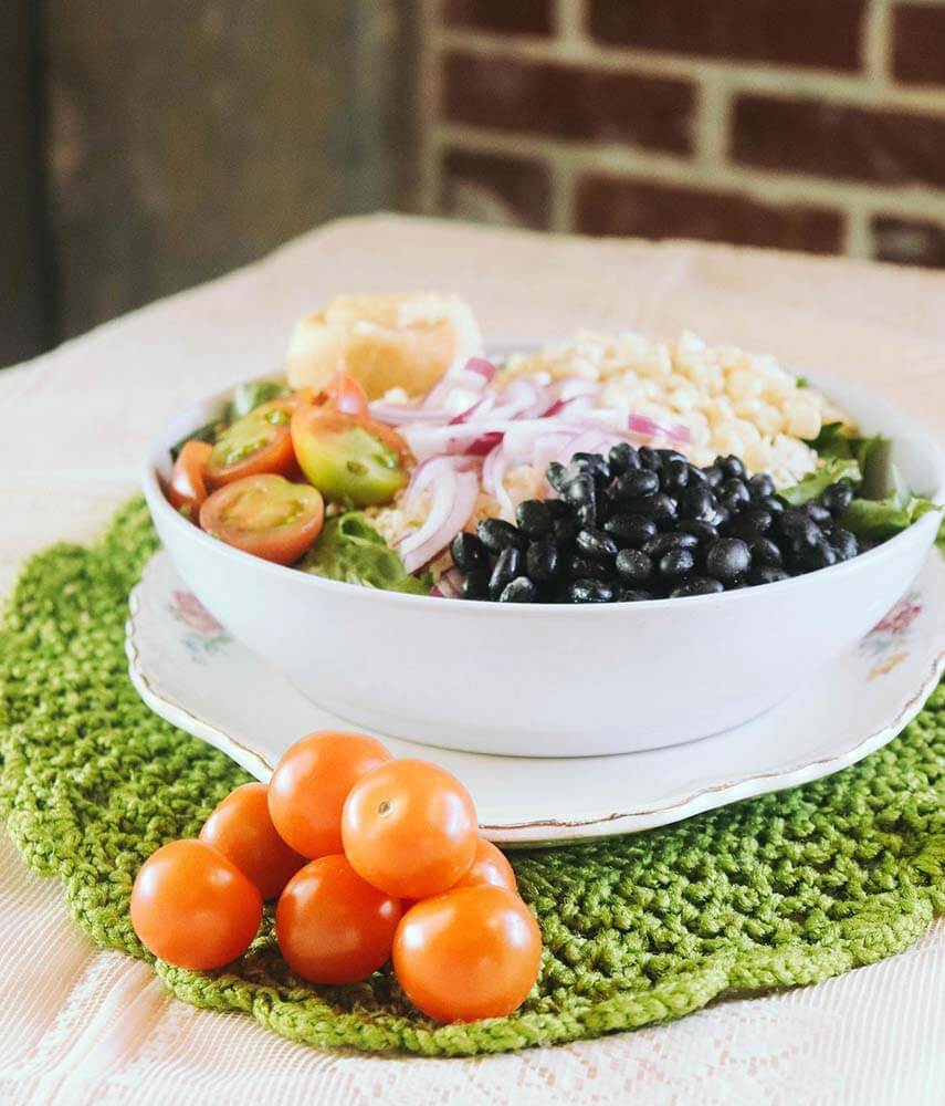 A white bowl filled with rice, black beans, sliced tomatoes, and red onions sits on a green mat. Whole cherry tomatoes are placed in front. A brick wall is in the background.