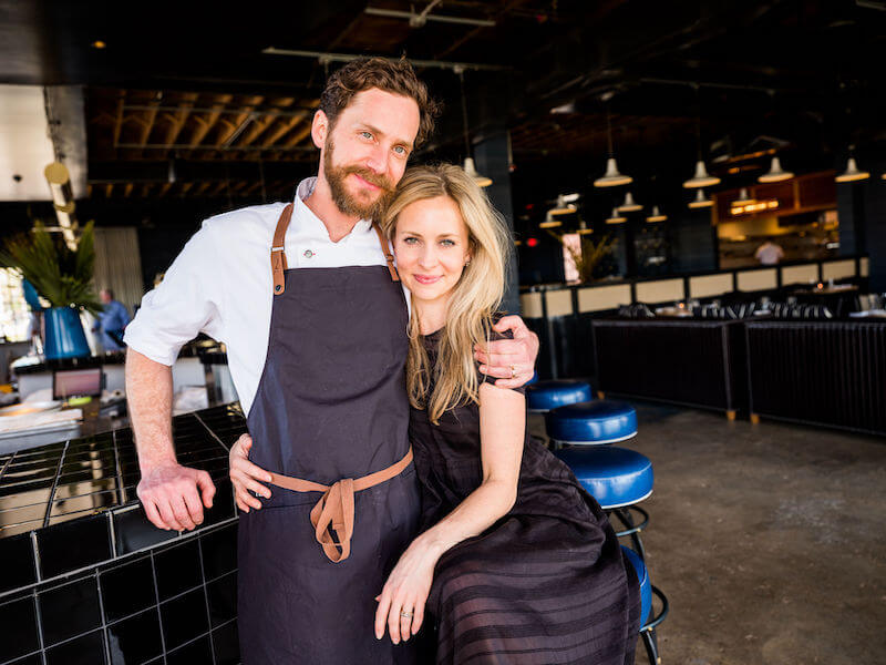 A man in a chef's outfit and a woman in a designer black dress are standing close together in a Birmingham restaurant, smiling at the camera.