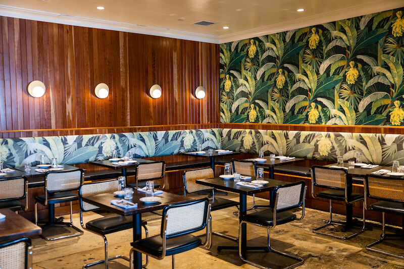 In a chic Birmingham restaurant, designer wooden paneling complements the tropical-themed wallpaper. Empty tables and chairs await guests, with place settings meticulously arranged for perfection.