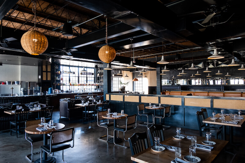 The interior of this modern Birmingham restaurant features designer wooden tables, dark chairs, and hanging spherical lights. Large windows and an open layout create an airy atmosphere.