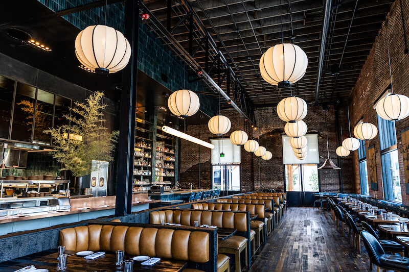 A modern restaurant interior in Birmingham features brick walls, designer spherical pendant lights, wooden floors, and leather seating. The open kitchen is visible, with neatly arranged tables providing a chic and welcoming dining experience.