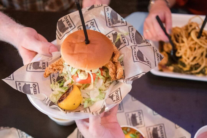 A person in Birmingham holds a popular chicken sandwich with lettuce and tomato, placed on branded paper, with a grilled lemon wedge on the side. In the background, another person enjoys pasta, setting the scene for a tasty 2024 dining experience.