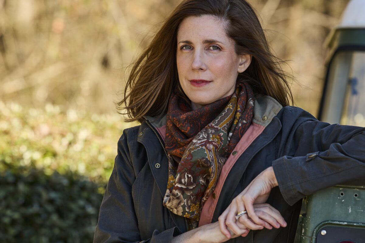 Mary Menge, co-owner of Tom Beckbe, wearing a jacket and patterned scarf, leans on a green vehicle outdoors, looking thoughtfully at the camera.