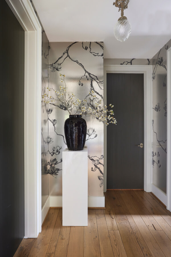 This hallway embodies future interior design trends for 2025, featuring a wooden floor, metallic floral wallpaper, and a dark door. A pedestal holds a black vase with branches beneath a sleek ceiling light fixture.