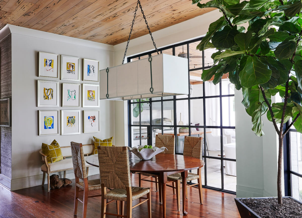 A dining room with a wooden table, wicker chairs, and a large leafy plant under a stylish pendant light captures 2025 interior design trends. The wall features framed artwork next to a windowed partition, blending elegance and modernity seamlessly.