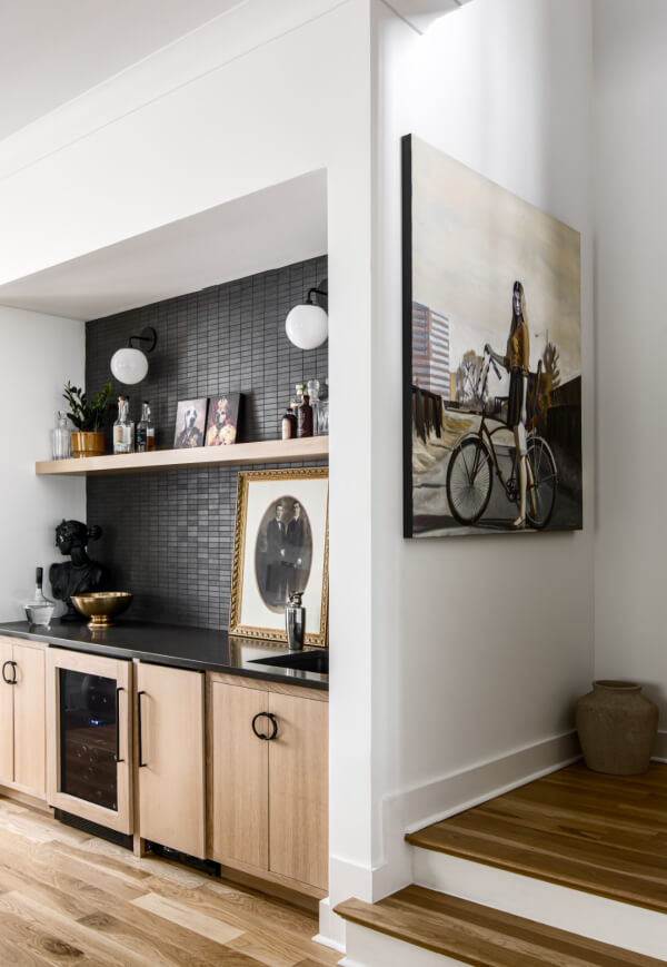 Modern kitchen with light wood cabinets, black countertop, and a stylish backsplash reflects 2025 interior design trends. Built-in shelves showcase framed photos and plants, while a large painting of a woman on a bicycle adds artistic flair.