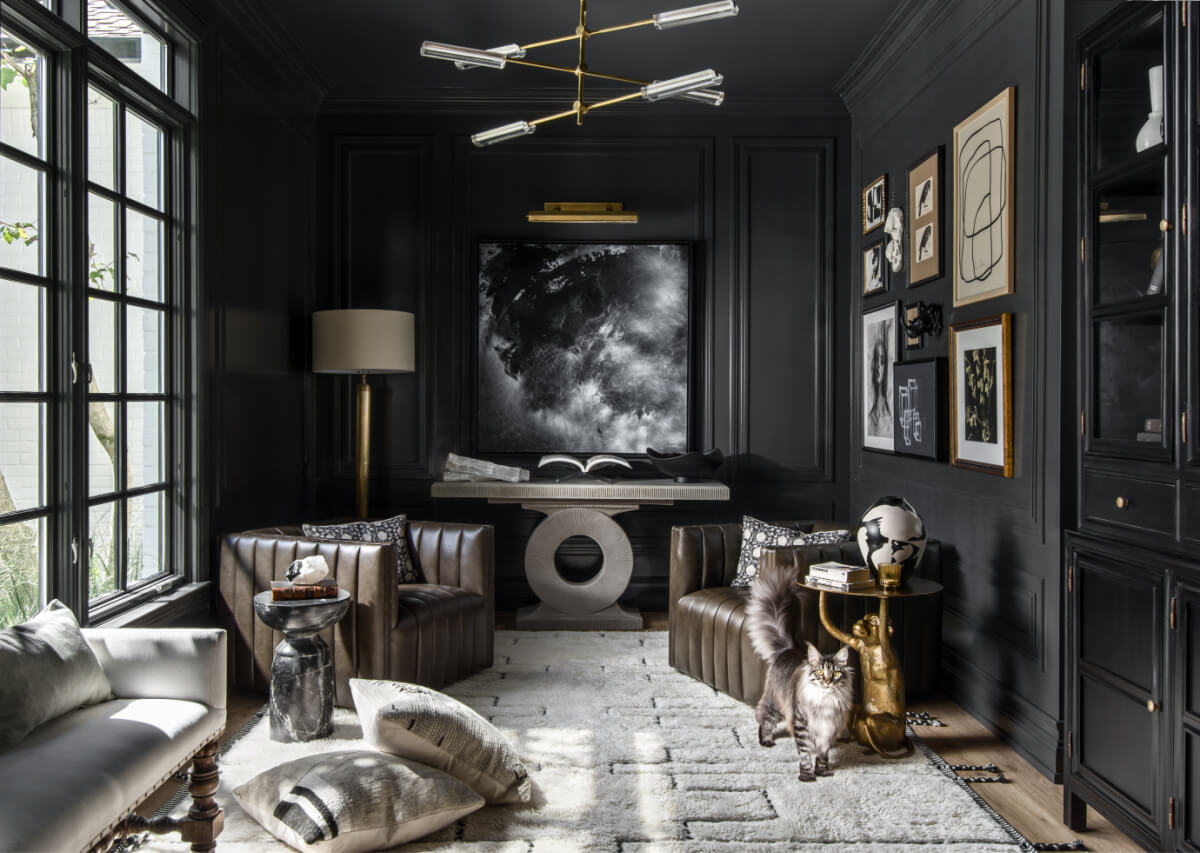 A modern living room featuring dark walls, leather chairs, and abstract art captures the essence of 2025 interior design trends. A cat strolls across a light-colored rug beneath a unique chandelier, while natural light floods through a large window.