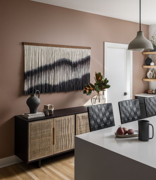 Discover a modern dining area that embodies 2025 interior design trends, featuring a sleek white island, black woven chairs, and chic pendant lighting. The walls showcase black-and-white abstract art, while shelves display decorative items and a vase bursting with lush foliage.