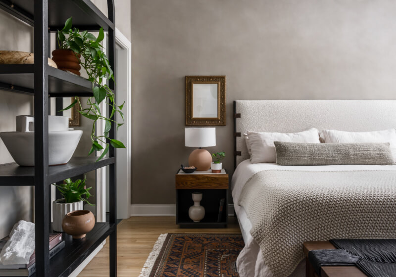 A minimalist bedroom features a textured gray wall and beige bedding, accented by a wooden side table. In line with 2025 interior design trends, a black shelf with plants and decor items stands to the side, while a framed mirror elegantly hangs above the nightstand.