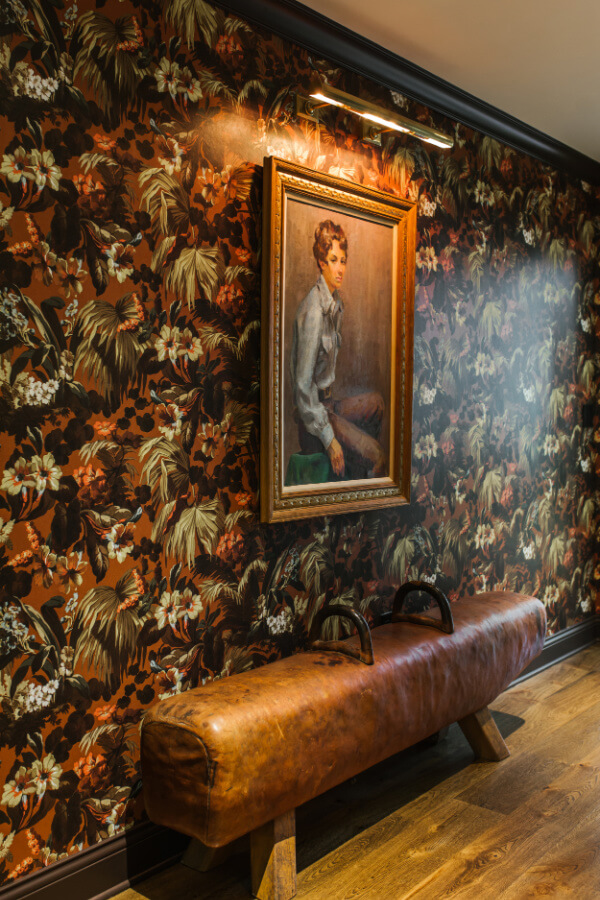 A framed portrait of a person hangs on a wall adorned with dark floral wallpaper, showcasing a nod to 2025 interior design trends. Below, a leather pommel horse bench elegantly rests on the wooden floor.