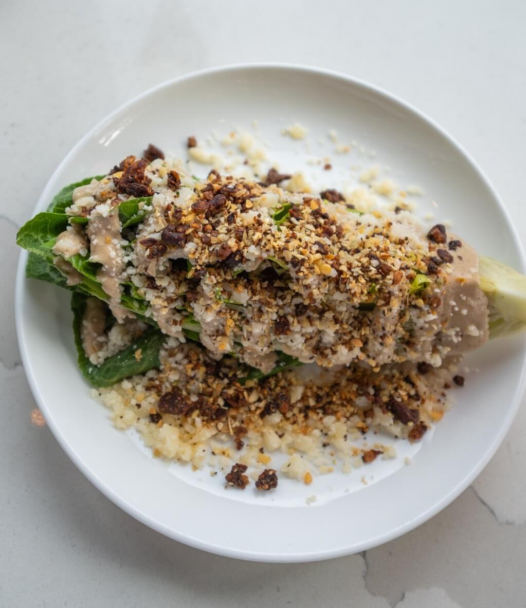 A wedge of lettuce topped with creamy dressing, crumbled cheese, and toasted breadcrumbs is served on a white plate, making it one of the best salads in Louisville.