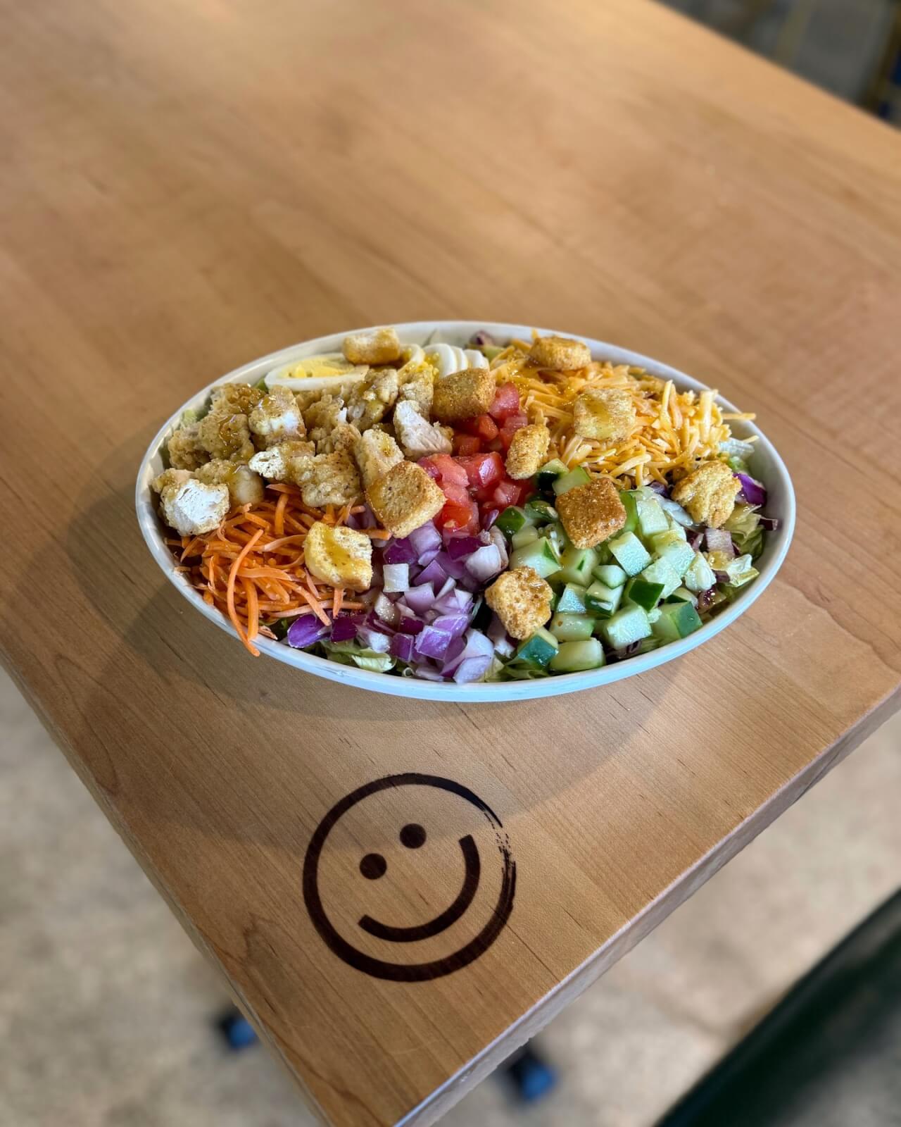 A salad bowl with croutons, shredded cheese, cucumbers, carrots, tomatoes, red onions, and various greens sits invitingly on a wooden table with a smiley face engraving—truly one of the best salads in Louisville.