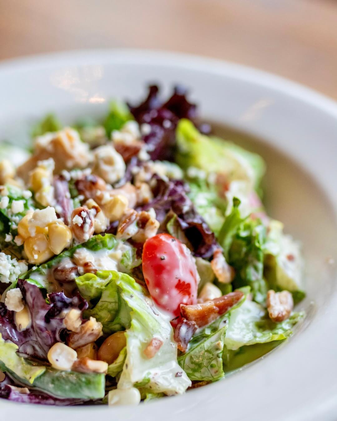 Bowl of mixed salad with lettuce, cherry tomatoes, cheese crumbles, nuts, and corn in a white dish.