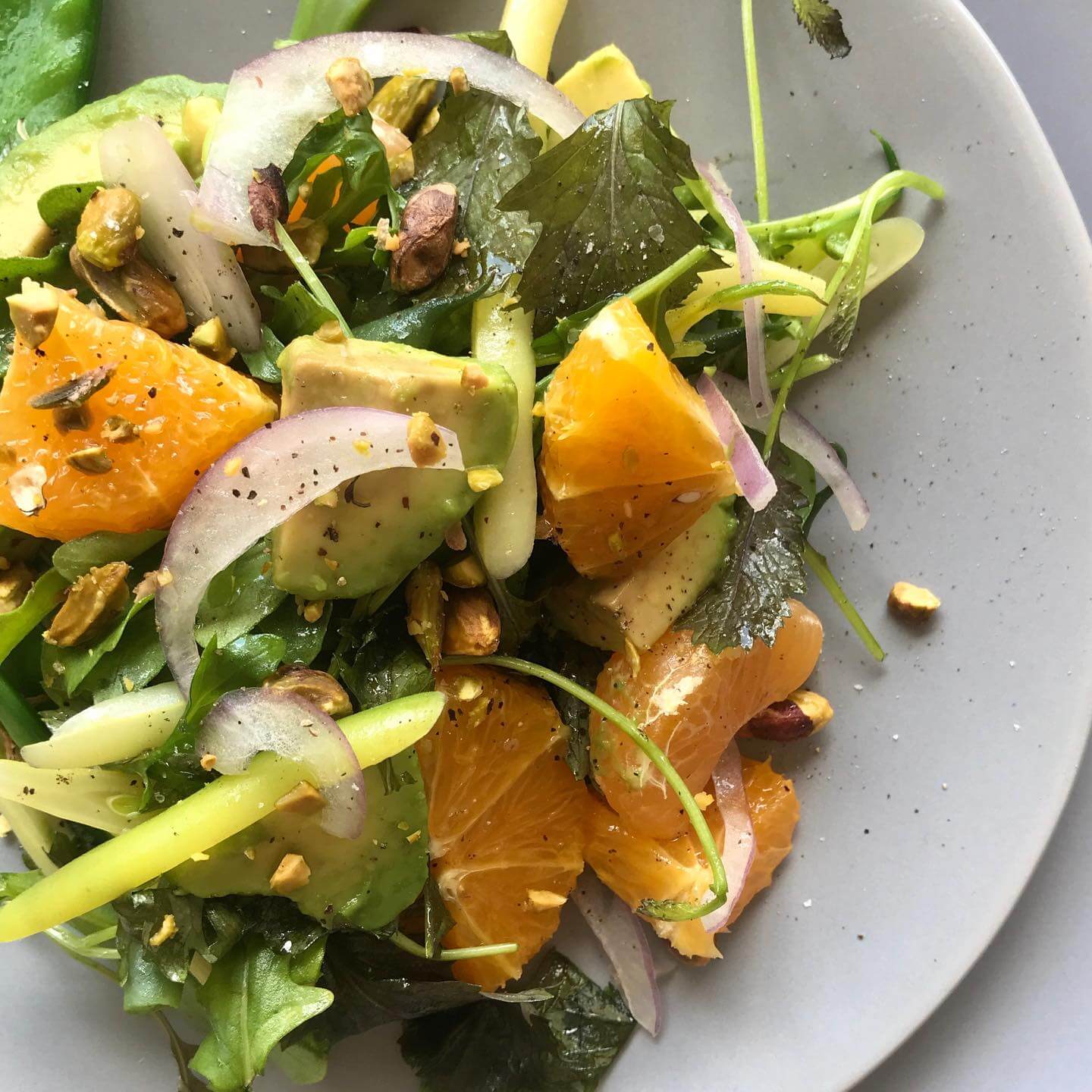 A fresh salad featuring sliced oranges, avocado, red onion, mixed greens, and pistachios elegantly arranged on a gray plate.