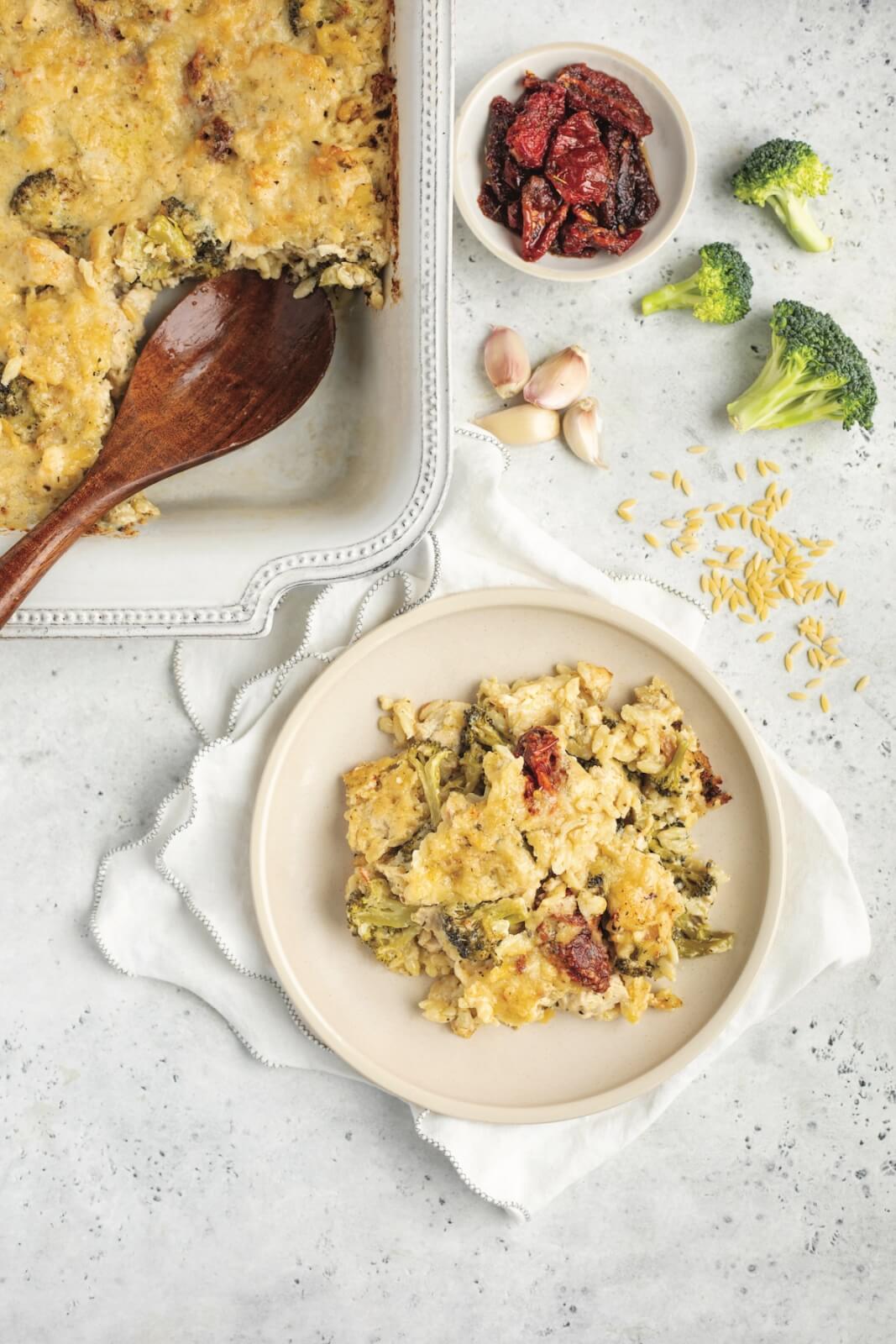 A plate of casserole with broccoli and sun-dried tomatoes, next to a casserole dish. Nearby are broccoli pieces, garlic cloves, orzo pasta, and a dish of sun-dried tomatoes on a napkin.