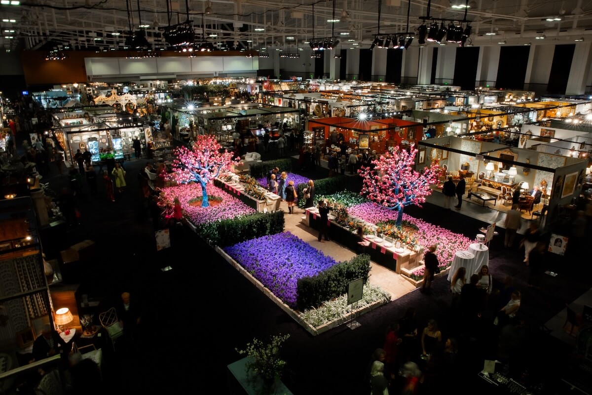 The Nashville Garden Show is a large indoor exhibition featuring colorful flowering trees and vibrant stalls with various products, including exquisite antiques, all under bright overhead lighting.