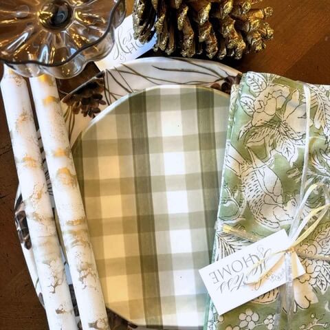 A green plaid plate, floral napkin, decorative wrapping paper, a glass dish, and a gold pinecone are arranged on a wooden table.