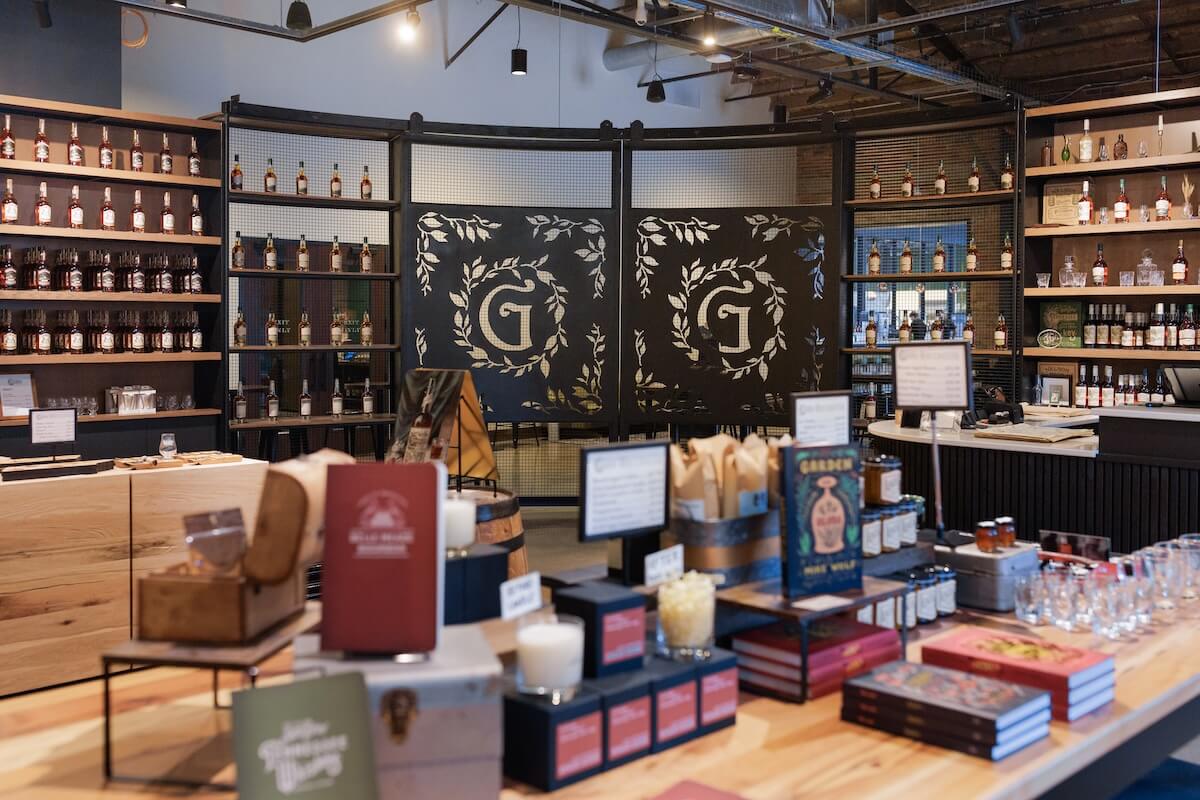 The charming store interior of Nelson’s Green Brier Distillery features shelves of bottles, a wooden counter adorned with packaged goods, books, and glassware. A decorative partition showcasing the "G" logo elegantly separates spaces, inviting you to explore the rich history and distinctive flavors within.