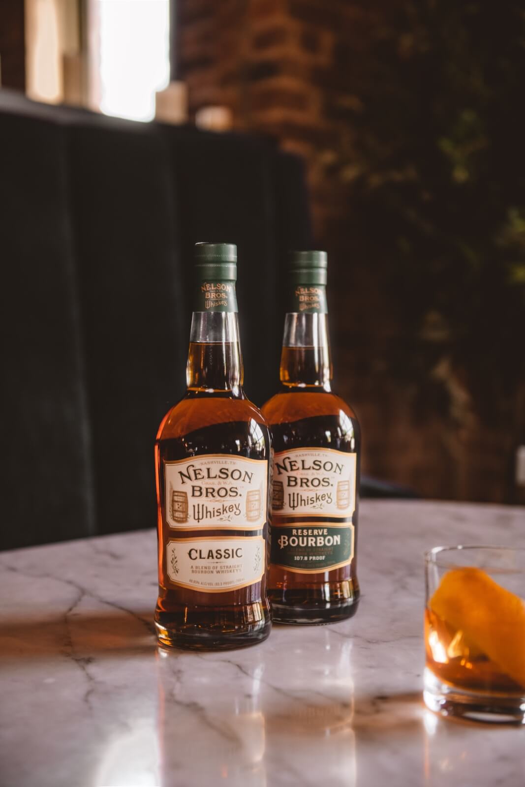 Two bottles of Nelson Bros Whiskey from the historic Green Brier Distillery stand on a marble table, one labeled "Classic" and the other "Reserve Bourbon." A glass with an amber drink and orange peel caters to those who appreciate rich flavor.