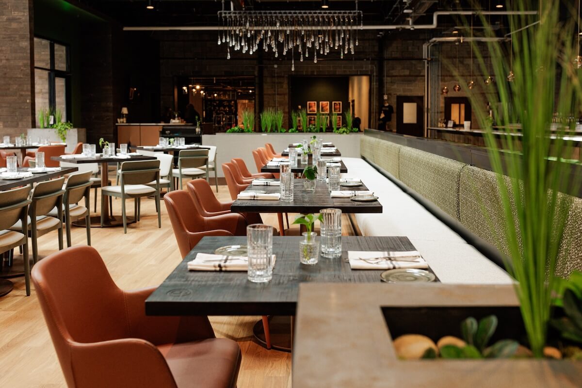 The modern restaurant interior features neatly arranged tables, leather chairs, and lush green plants, illuminated by a ceiling chandelier that echoes the innovative spirit of Nelson’s Green Brier Distillery.