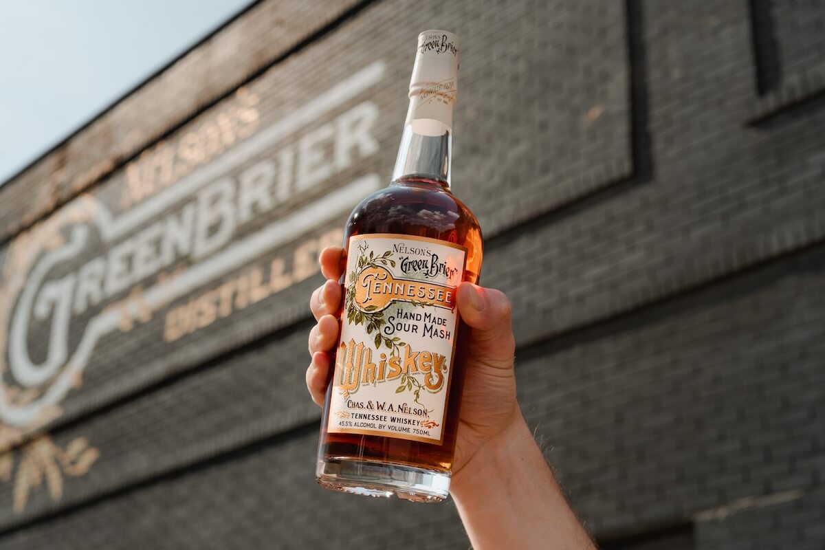 A hand holds up a bottle of Green Brier Tennessee whiskey against a brick wall, with hints of the brand’s historic signage visible. The rich flavor carries the legacy of Nelson’s Green Brier Distillery in every sip.