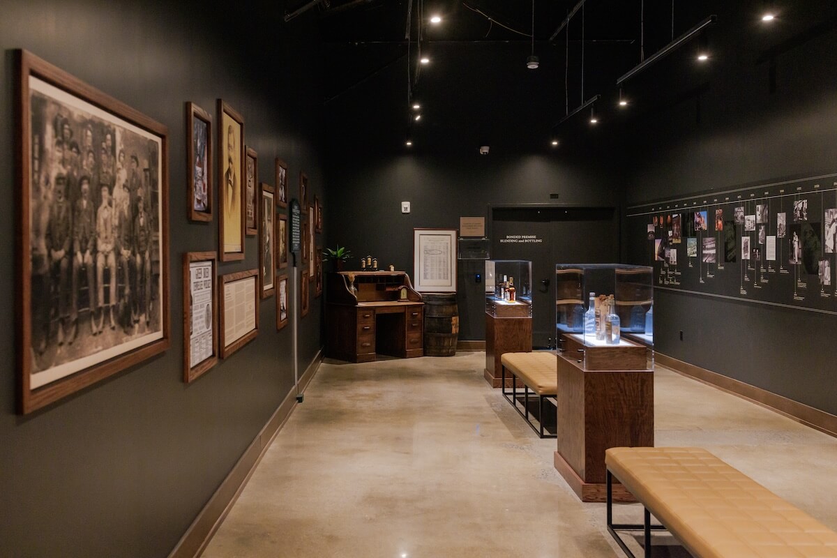 The exhibit at Green Brier Distillery showcases framed photos and historical documents on black walls, with a wood desk and display cases. Benches line the center aisle, inviting guests to sit and reflect on the rich history that drove innovation in distilling.