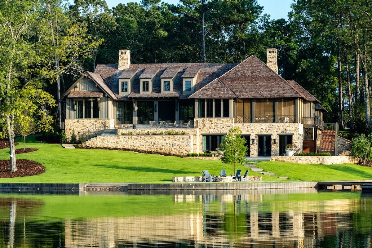 A stone and wood house with a sloped roof sits by a calm lake, nestled in one of the serene second home communities, surrounded by lush greenery and trees under a clear sky.