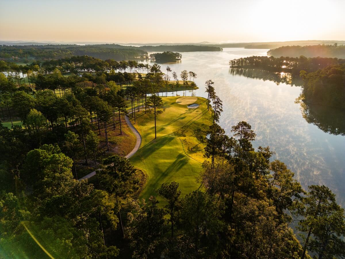 Aerial view of a golf course in serene second home communities, with a green surrounded by trees and a nearby lake, all illuminated by the gentle light of sunrise.