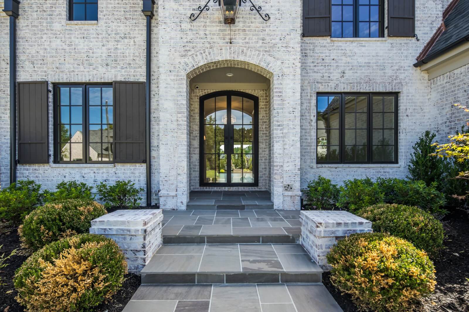 The front entrance of the brick house showcases an arched glass door, flanked by large windows and surrounded by neatly trimmed bushes. Steps adorned with natural stone lead up to the inviting doorway.