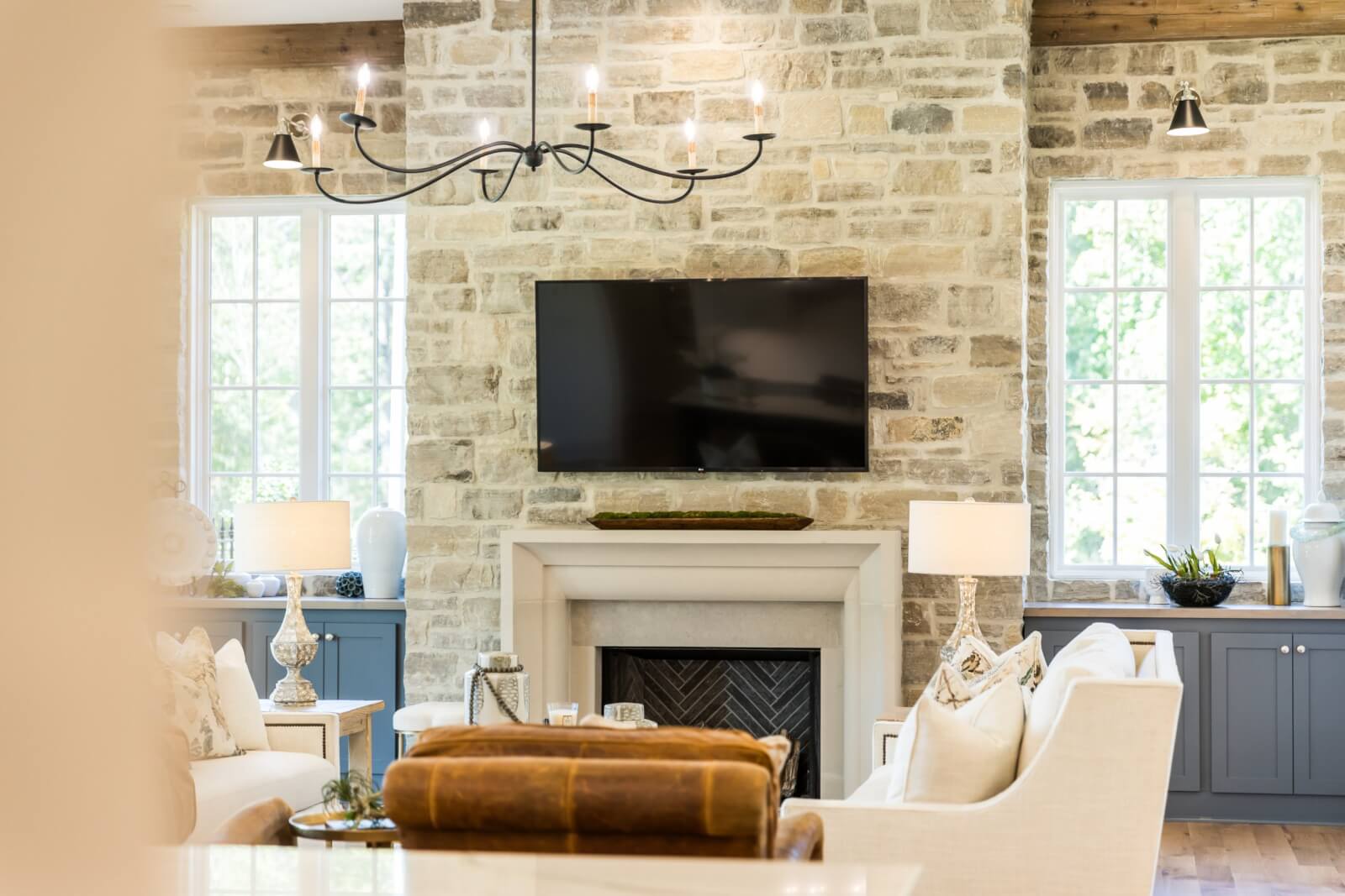 This stylish living room features a natural stone fireplace, a wall-mounted TV above, and two windows that illuminate the space. Blue cabinets contrast with cream sofas, while decorative items add charm beneath a striking black chandelier, creating an inviting home atmosphere.