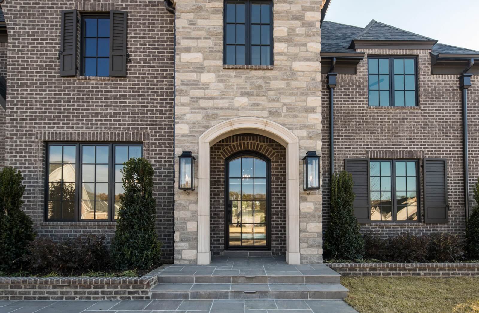 Front view of a modern brick house with a stone arch entrance, large windows, and black shutters, incorporating natural stone elements. The home decor is enhanced by small shrubs surrounding the facade.