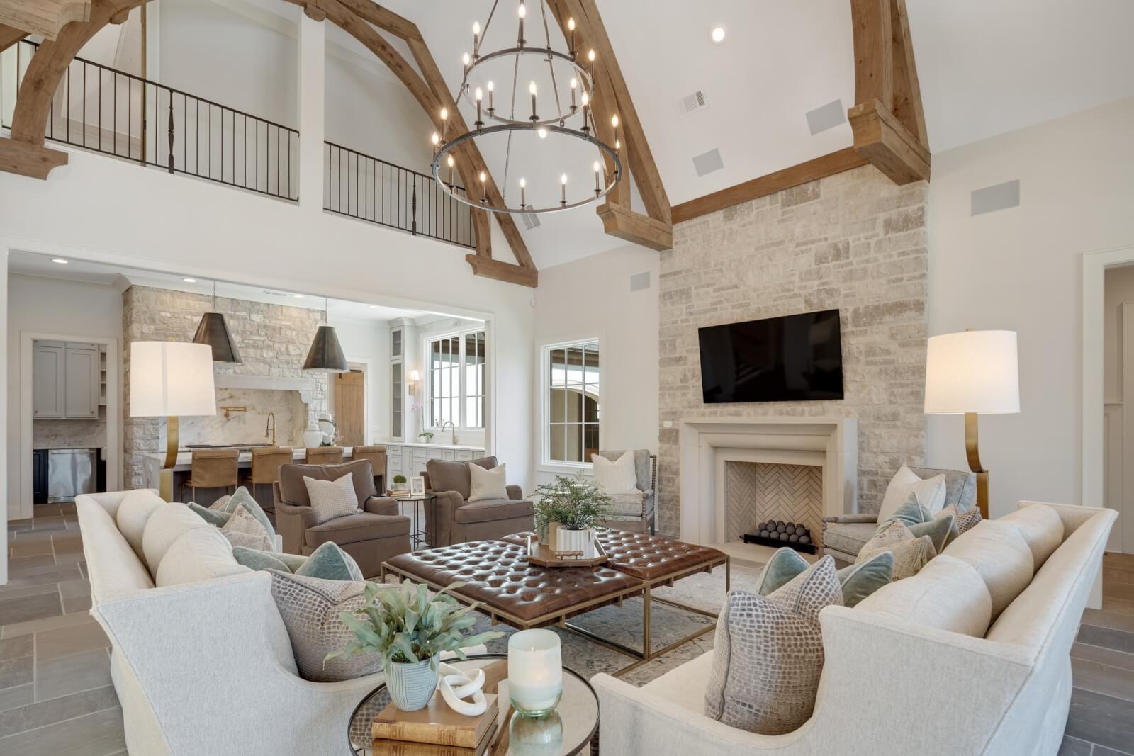 Spacious living room with high wooden beam ceiling, natural stone fireplace, chandelier, and neutral-colored sofas arranged around a central coffee table. Large windows illuminate the space, seamlessly incorporating elegant home decor elements.