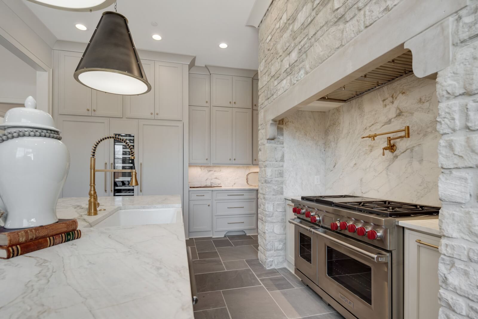 This modern kitchen features gray cabinets, marble countertops, and a large stove with red knobs. The tiled floor pairs beautifully with the stone accent wall, incorporating natural stone elements. Gold-toned faucet and large pendant lights enhance the sophisticated home decor.