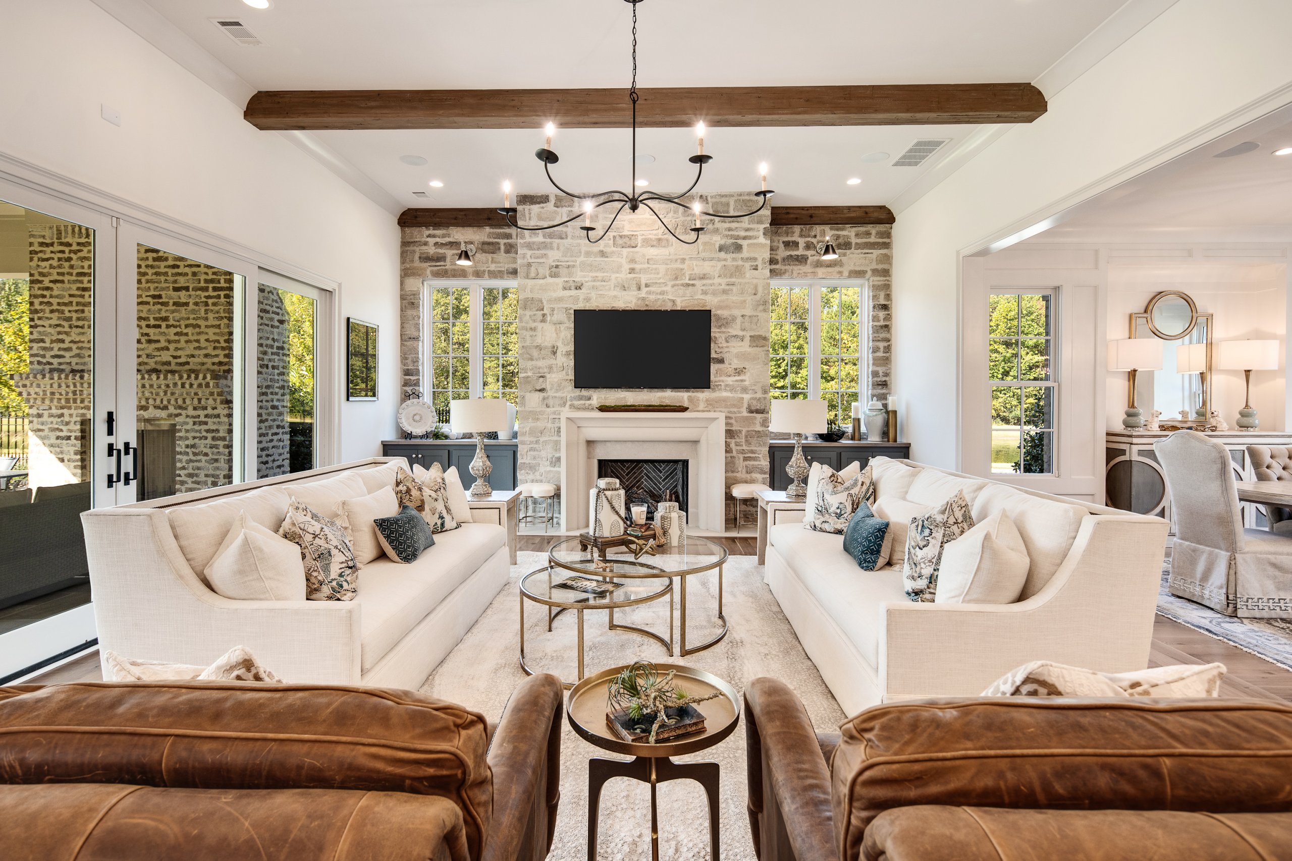 Elegant living room with two cream sofas and a stone fireplace that beautifully incorporates natural stone. Wooden ceiling beams and a chandelier add charm, while large windows and glass doors provide natural light, leading to an outdoor view. Perfect for those who appreciate refined home decor.