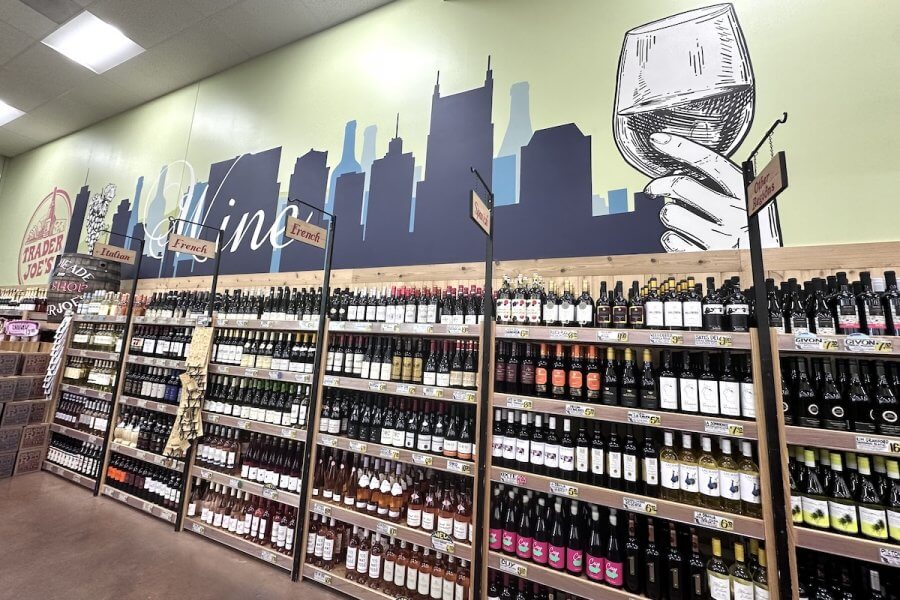 A store aisle displays a variety of Trader Joe's wines on shelves beneath a mural featuring a city skyline and a hand holding a wine glass.