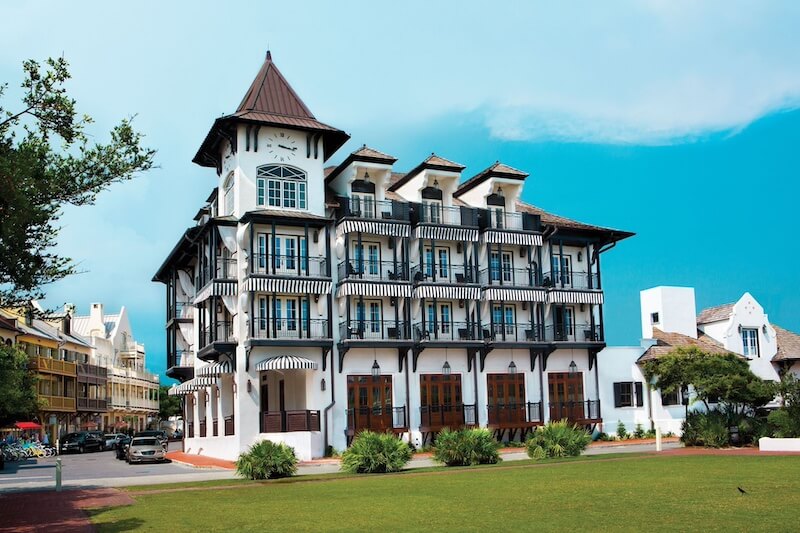 Luxurious multi-story white building with black balconies, turret, and coastal 30a architectural style, situated near a green lawn under a blue sky.