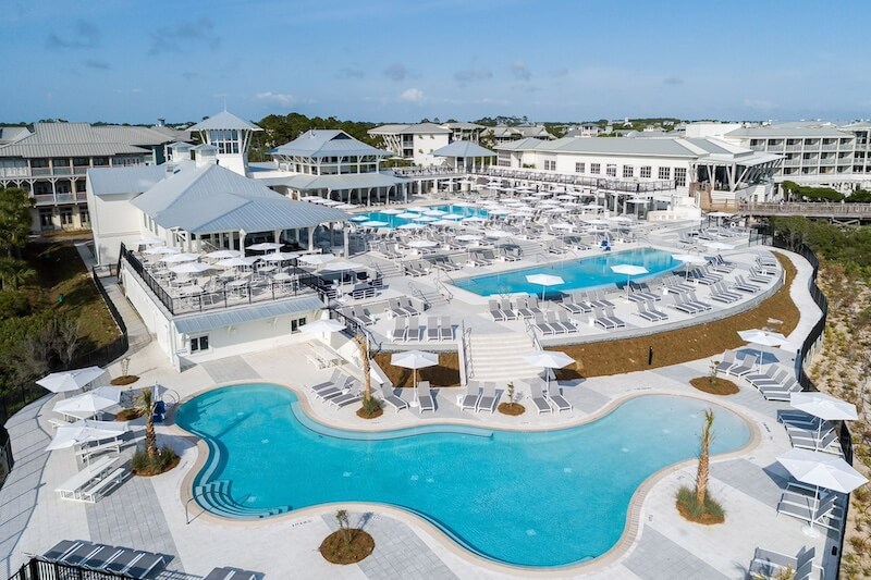 Aerial view of a 30a resort with multiple swimming pools, loungers, and a central building with a white roof, surrounded by smaller white buildings.