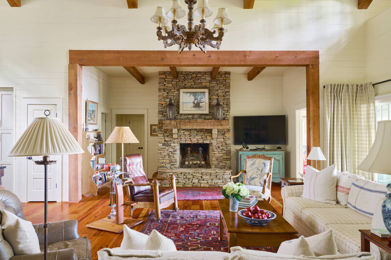 A cozy living room in a Lake Martin cabin features a stone fireplace, a chandelier, wooden beams, white sofas, wooden armchairs, various lamps, a television on the right, and decorative rugs on a hardwood floor.