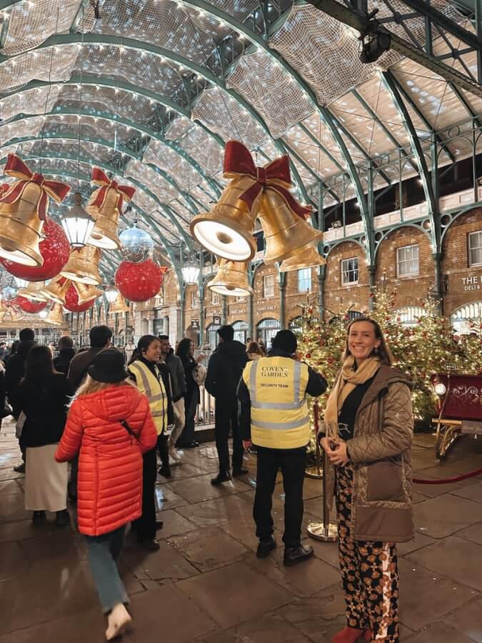 People strolling under festive holiday decorations in a bustling indoor venue. It's a lively scene during the long weekend in London, with a person in a yellow vest at the center and another in a brown coat on the right, smiling warmly at the camera.