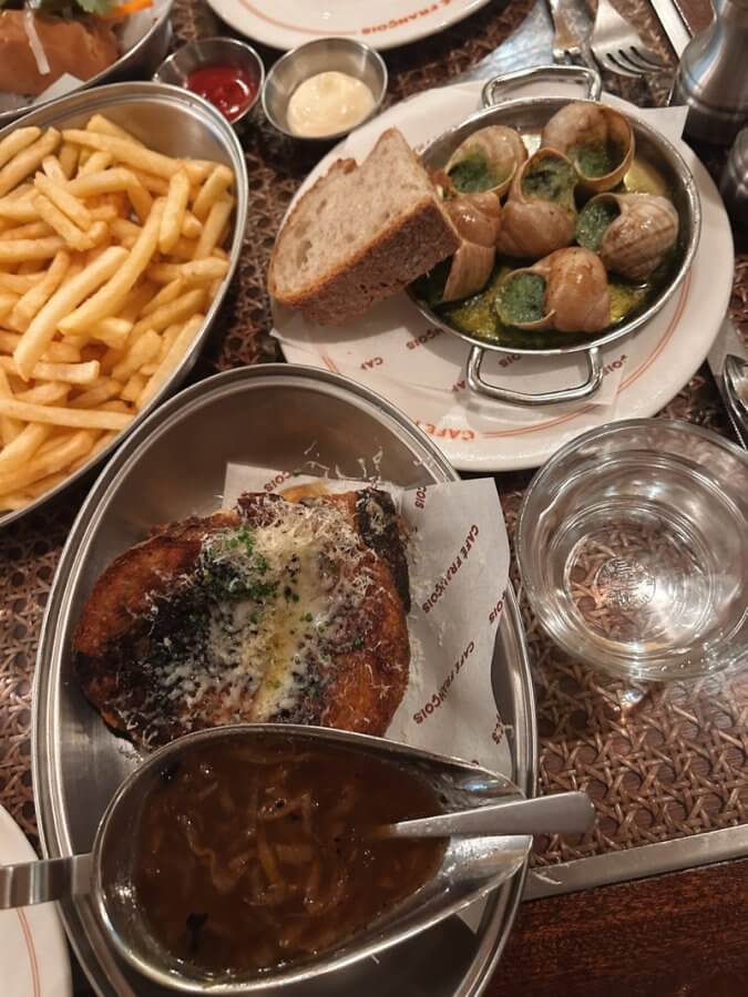 Amidst dishes like escargot with bread, French fries, and a plate of French onion soup, the table hints at indulgence during a long weekend in London. A glass of water sits elegantly beside these delights.