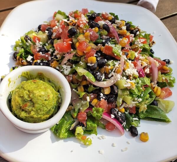 A colorful salad with lettuce, black beans, corn, tomatoes, onions, and cheese graces a white plate alongside a small bowl of guacamole. It's one of the best salads in Memphis for its vibrant flavors and fresh ingredients.